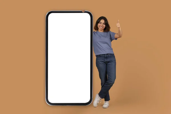 素晴らしいオンラインオファー 肯定的な若い魅力的なアラブの女性は広告のための白い空白のスクリーンのモックアップのコピー スペースが付いている大きい電話によって立って 親指を示します 多彩な背景 — ストック写真