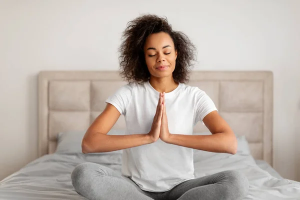 stock image Calm black young woman meditating in lotus position with hands clasped together, relaxing during morning yoga, sitting on bed at home bedroom interior, embracing mindfulness and relaxation