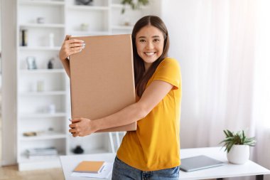 Neşeli, genç Asyalı bir kadın elinde karton bir kutu tutarken masasının yanında, mutlu Koreli bir kadın boks yapmaya hazır, hızlı teslimat hizmetlerinden hoşlanıyor, kameraya gülümsüyor, uzay boşluğunu kopyalıyor.