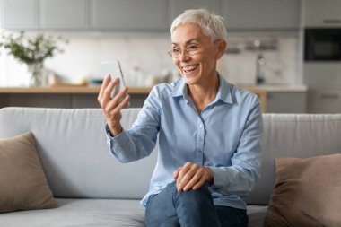 Son sınıf öğrencisi neşeli bir kadın cep telefonunu tutuyor ve kullanıyor. Gözlük takıyor, internette sohbet ediyor, mesajlaşıyor, web sitesinde geziniyor. Modern ev içlerinde rahat bir kanepede oturuyor.