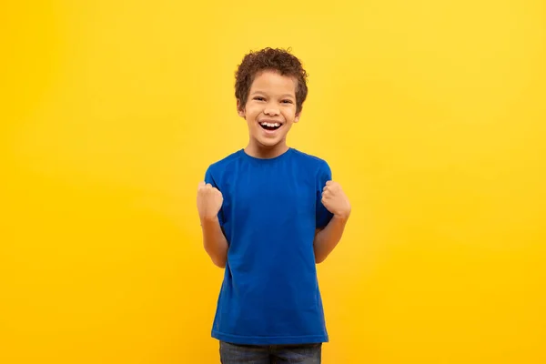 カーリーな髪の男の子は笑い 勝利で拳をクレンジング 成功を祝い 黄色の背景に対して王室の青のTシャツを着ています — ストック写真