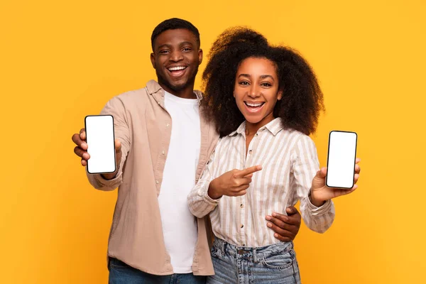 Smiling young black man and woman happily present smartphones with white blank screens, pointing at them, ready for digital branding on yellow background