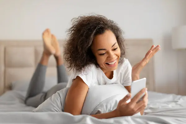 Joyful Black Millennial Lady Communicating Via Cellphone, Making Video Calls And Reading Great News Online, Lying In Bed At Home Interior. Excited Black Woman Using Mobile Application On Phone