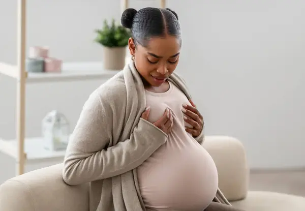 忧心忡忡的年轻怀孕的非洲裔美国女人 胸部有痛苦的感觉 按摩自己 坐在沙发上 自由自在 未来的母亲保健 — 图库照片