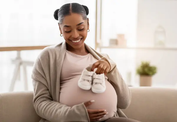 等待一个孩子 快乐而美丽的年轻非洲裔美国孕妇坐在家里的沙发上 在她的大肚子上抱着可爱的小婴儿靴 微笑着 复制空间 — 图库照片