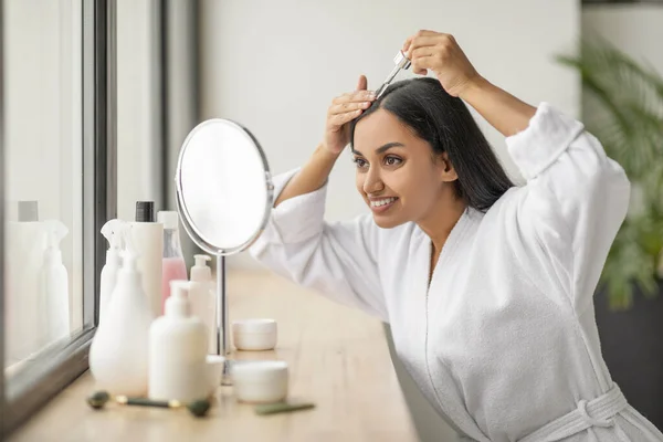 Positive pretty young indian woman wearing white bathrobe looking at mirror, using nourishing hair oil for hair growth, dropping serum on scalp. Hair care at home