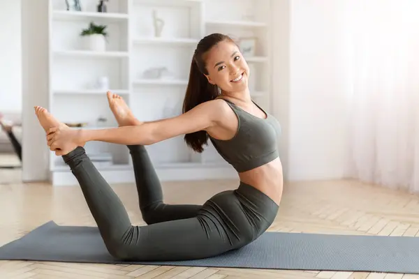 适合年轻的亚洲女人在家里摆出富有挑战性的瑜伽姿势 对着镜头微笑 快乐而活泼的韩国女人展示力量和灵活性 在宽敞的客厅里做健身垫的训练 — 图库照片