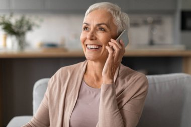 Olumlu, olgun bir bayanın hoş bir telefon konuşması gülümsemesi, kulağına akıllı telefon dayaması, modern ev içinde mobil iletişimin keyfini çıkarması, portre çekimi.