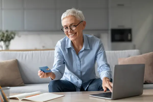 Ecommerce. Smiling mature woman shopping online making financial transaction via debit credit card and laptop computer, sitting on comfortable sofa at home indoors, wearing eyeglasses