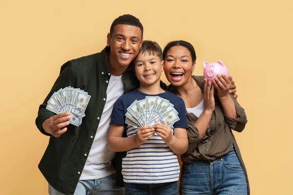 Family Money. Happy Family Father Mother And Son Showing Piggybank And Paper Notes Dollar Cash To Camera Raising Savings Together, Beige Studio Background. Great Financial Offer Concept