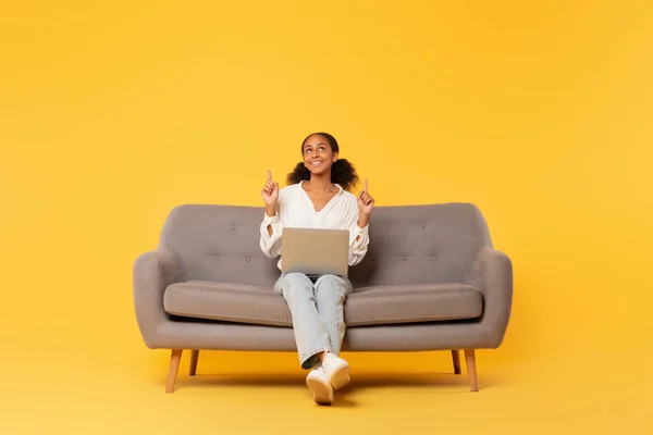 Happy African American teenager girl studying on her laptop, pointing fingers up in moment of eureka, sitting casually on sofa against yellow studio backdrop. Student having idea during learning