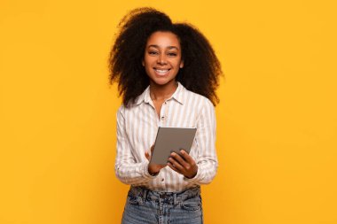 Kıvırcık saçlı, çizgili gömlekli, kendinden emin genç bir kadın tablet taşıyor. Sıcak gülüşü, güneşli sarı arka planda verimlilik olduğunu gösteriyor.