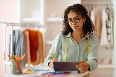 Modern stüdyosundaki canlı renk örnekleriyle çevrili, tablette stilus ile dijital olarak çizim yapan konsantre genç siyahi kadın moda tasarımcısı.
