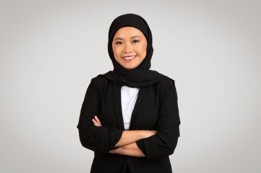 Siyah tesettürlü ve ceketli kendine güvenen Müslüman iş kadını kollarını kavuşturmuş, profesyonel bir ortamda gülümsüyor, liderlik ve güven yayıyor, gri arka planda, stüdyoda izole edilmiş.
