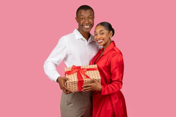 赤いリボンと包まれたギフトボックスを保持しているハッピーブラックカップル 赤いシャツの男性と赤いドレスの女性両方の笑顔 ピンクの背景 — ストック写真
