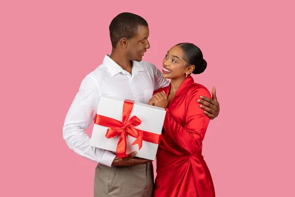 Delighted black man in white shirt presents gift with red ribbon to appreciative woman in red dress, sharing joyful moment of giving on pink background