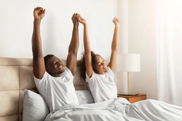 Happy young african american couple satisfied after good sleep raising and stretching arms, sitting together in bed in their modern bedroom interior, enjoying morning. Awakening on weekend