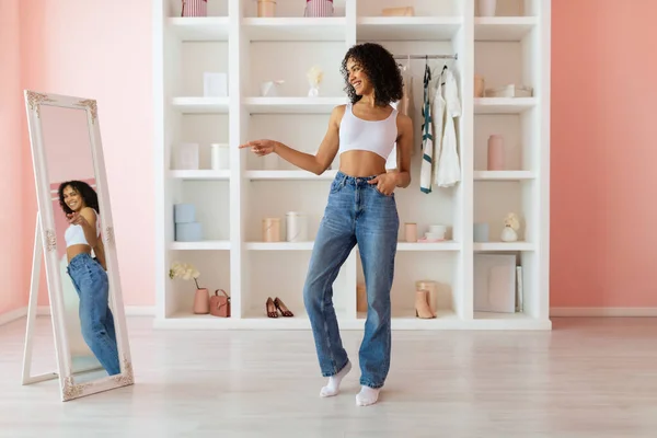 Cheerful young woman in casual jeans and white top, pointing with joy at her reflection in full-length mirror in a bright, cozy room interior
