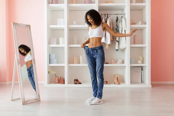 Curly-haired latin woman proudly showing her weight loss success by wearing too-large jeans, reflected in a full-length mirror in a light room