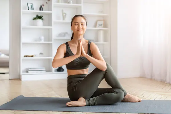 Serene asian woman practicing yoga in twist pose at home, calm korean female sitting on fitness mat and keeping hands in namaste gesture, exuding tranquility in well-organized living space