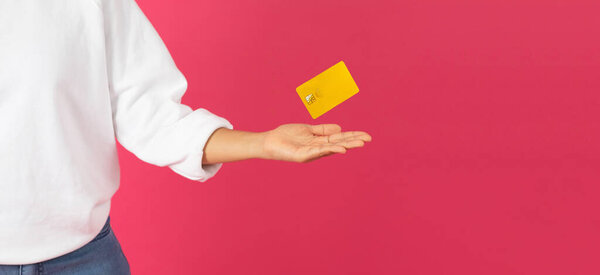 Magical Finance. Young woman levitating yellow credit card with her palm, unrecognizable female recommending bank services, isolated against vibrant pink background, panorama with copy space