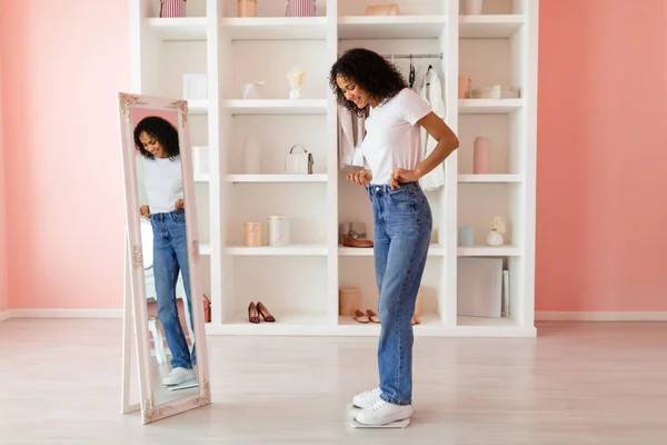 Young latin woman in casual attire happily admiring her fit in blue jeans and white t-shirt, reflected in full-length mirror in a stylish room
