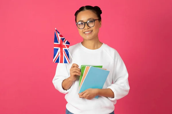 美丽的年轻亚洲女学生举着英国国旗和书本 快乐的韩国女学生在明亮的粉色背景下推荐国际学习和交流项目 — 图库照片