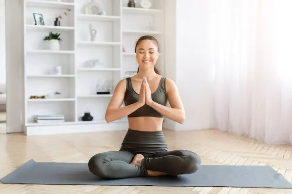 Wellness Concept. Serene young asian woman in yoga pose, meditating with eyes closed, beautiful korean female sitting on fitness mat with clasped hands, practicing meditation at home