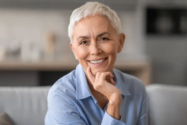 Aged Beauty. Portrait of relaxed gray mature lady with short haircut sitting on the couch indoor in living room and posing. Positive older woman looking and smiling at the camera