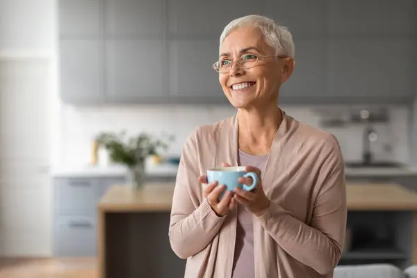 Happy mature woman with stylish haircut holds her tea cup posing in eyeglasses, standing in modern kitchen, embodying relaxed retirement life and happiness. Morning coffee. Copy space