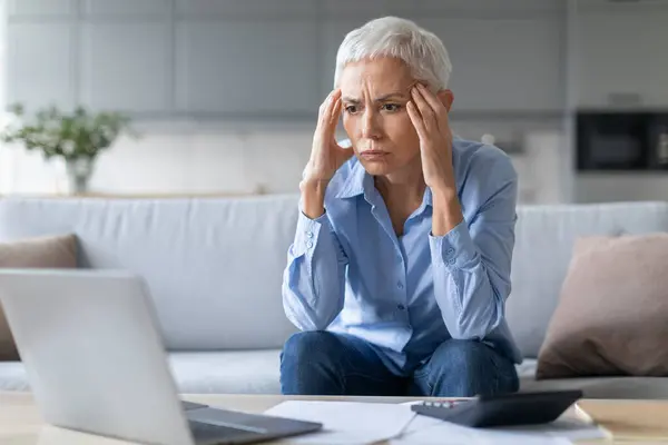 Stressed Mature Woman Touching Head In Despair While Calculating Her Budget And Expenses During Financial Crisis, Using Laptop While Doing Paperwork At Home. Bankruptcy Concept