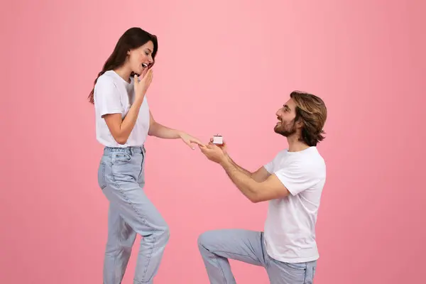 Happy young caucasian man on knee give ring box to surprised woman, isolated on pink background, studio. Surprise for anniversary, Valentine day and proposal at romantic date