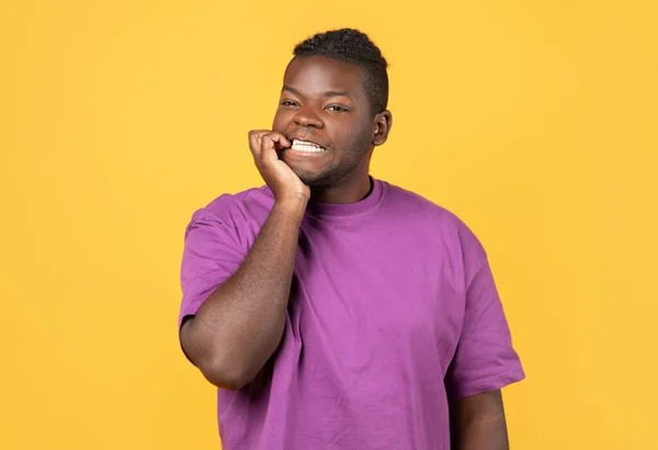 身穿紫色T恤的非洲裔美国人 穿着黄色背景的T恤 一边咬指甲一边表达焦虑 紧张和压抑情绪的概念 对紧张情况的反应 — 图库照片