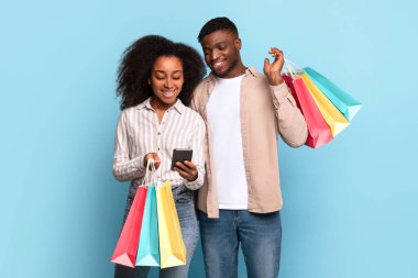 Genç siyah çift akıllı telefonla meşgul, kadın kaydırma ekranı, renkli alışveriş torbaları tutan adam, her ikisi de mavi arkaplanda dijital anı paylaşıyor.