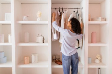 Curly-haired woman in casual attire thoughtfully arranging her stylish clothing in an open white closet in a room with soft pink walls clipart