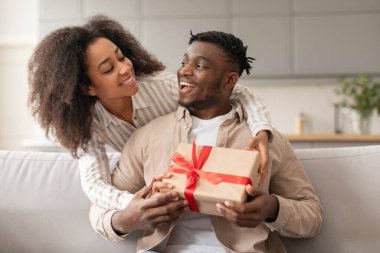 Sevgililer Günü hediyesi. Neşeli siyah eş, kocasının modern oturma odasının içinde ona hediye paketi vererek romantik bayramı veya ilişki yıldönümünü kutluyor.