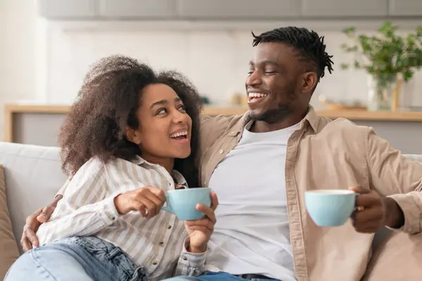 Cheerful Black Millennial Couple Drinking Morning Coffee Relaxing On Sofa At Home, Happy Romantic African American Young Spouses Enjoying Hot Drinks, Embracing Holding Cups