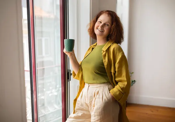 街の建築を眺めながら自宅の窓に緑のマグカップを掲げる陽気なシニア女性 明るいインテリアで平和な瞬間を楽しむ幸せな高齢女性 引退時にリラックス — ストック写真