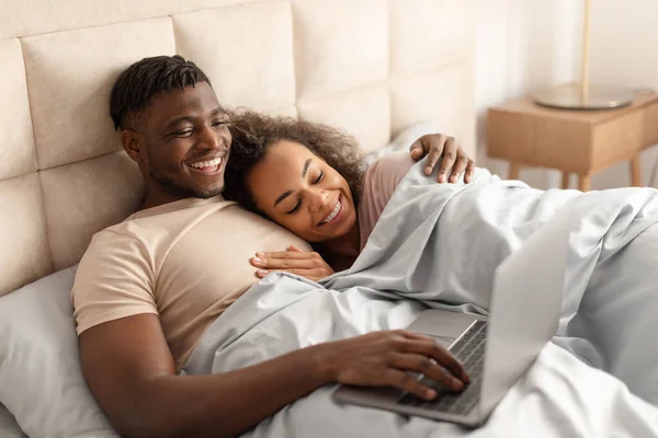 Happy black man and woman in love using laptop while lying in bed, spending weekend together at home bedroom, surfing web and watching funny videos online, hugging in modern bedroom