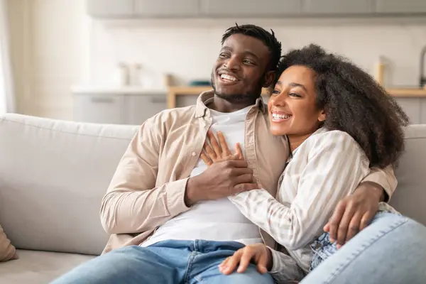 Marriage And Relationship. Cheerful African American Lovers Couple Cuddling Relaxing On Couch, Sharing Warm Embrace, Looking Aside With Smile, Resting In Modern Living Room, Free Space