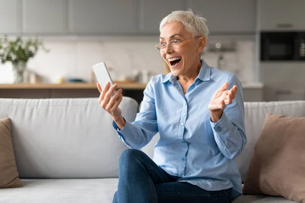 Wow mobile offer. Emotional mature woman looking at cellphone joyfully and gesturing, celebrating while reading amazing message online, sitting on sofa at modern living room indoor