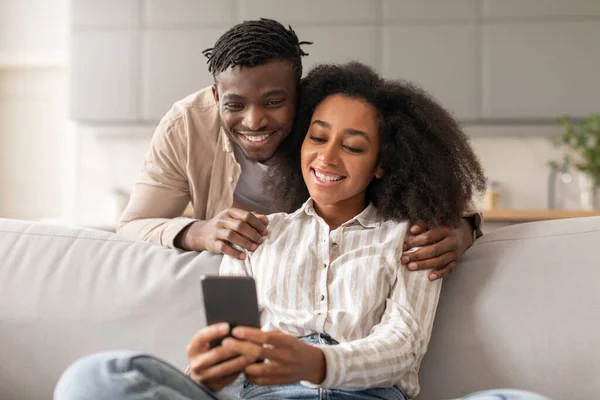 Cheerful black couple sharing social media on cellphone, happy african american family sitting on couch, using smartphone at home indoors, man embracing his wife looking at mobile phone screen