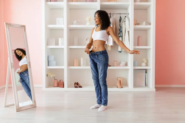 快乐的年轻女子穿着宽松的蓝色牛仔裤 在一个明亮的粉色调房间里 在镜子前展示她减肥的成功 — 图库照片