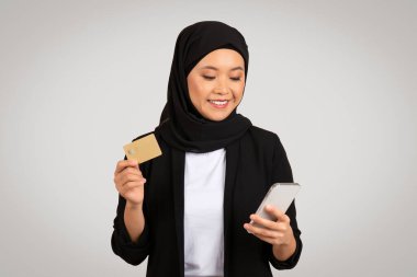 Türbanlı, ceketli ve beyaz gömlekli neşeli bir Müslüman kadın bir elinde akıllı bir telefon, diğer elinde altın bir kredi kartı tutuyor.