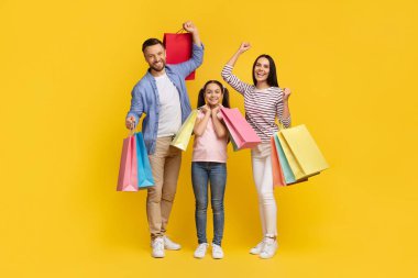 Mevsimlik satışlar. Bir sürü parlak alışveriş çantası tutan mutlu beyaz aile portresi, neşeli genç ebeveynler ve stüdyoda sarı arka planda poz veren kadın çocuk, kopyalama alanı.
