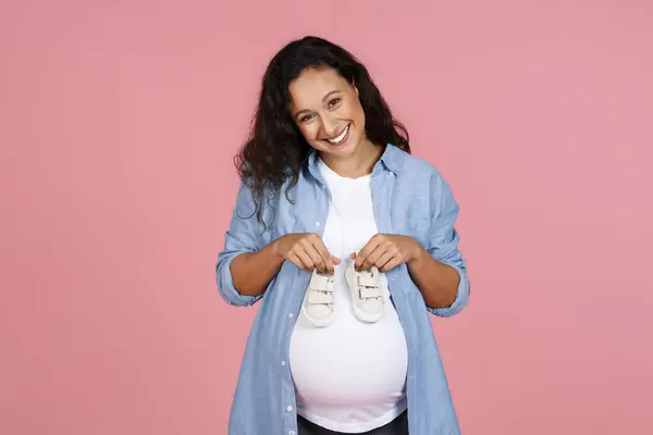 Waiting for a child, pregnancy. Joyful beautiful pregnant young woman holding cute little baby booties on her big tummy and smiling, copy space, isolated on pink studio background