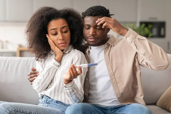 Unintended pregnancy. Concerned black spouses upset with positive test result not ready to be parents, sitting on sofa shocked and unhappy, posing at home. Unplanned Childbirth concept