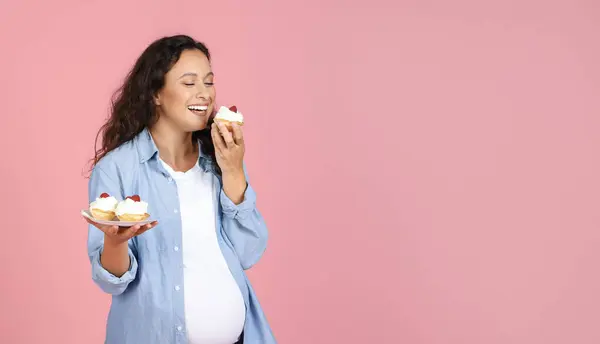 Hungry Expectant Lady Eating Sweets Standing Over Pink Studio Background. Foods to Avoid During Pregnancy Concept. Panorama, Empty Space