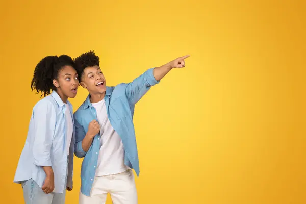 Excited African American young man pointing away with a thrilled expression as the young woman next to him looks on with surprise and interest, against a vivid yellow background