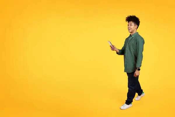 Chill stylish young african american guy with smartphone in his hand walking towards copy blank space for advertisement, isolated on yellow studio background